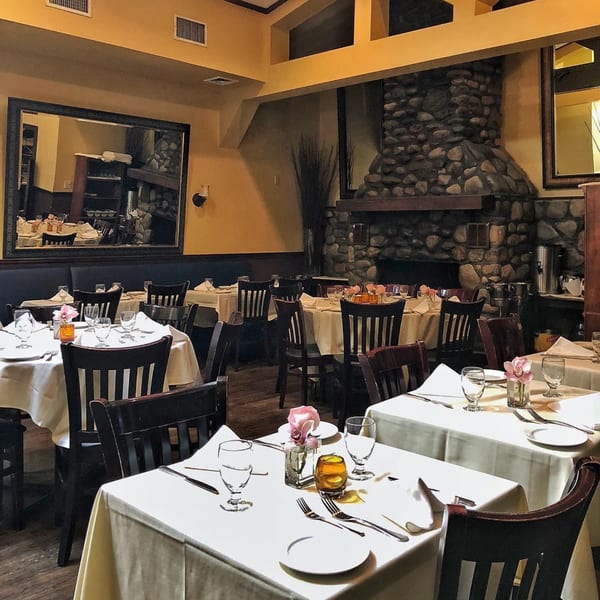 Elegant Italian restaurant interior with set tables, dark wooden chairs, and a large stone fireplace.