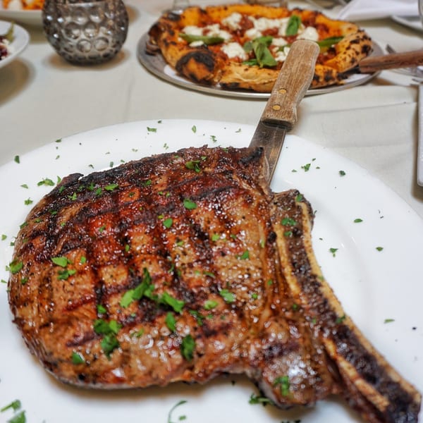 Grilled steak with a knife on a white plate, with a pizza in the background at Da Nicola.