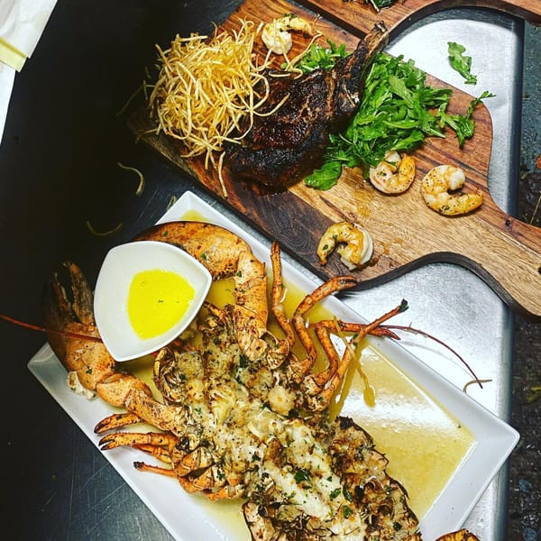 Grilled lobster with butter sauce and a steak with fries, shrimp, and arugula on a wooden board.