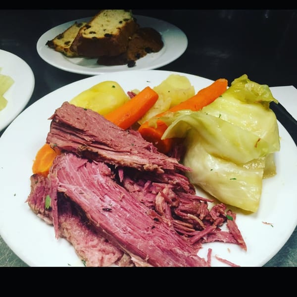 A plate of corned beef with boiled potatoes, carrots, and cabbage, with raisin bread in the background.
