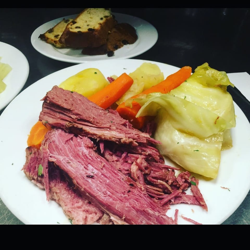 A plate of corned beef with boiled potatoes, carrots, and cabbage, with raisin bread in the background.