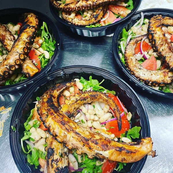 Four black bowls of grilled octopus salad with arugula, cannellini beans, tomatoes, and red onion.
