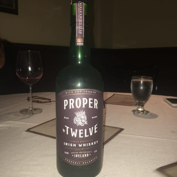 A bottle of Proper Twelve Irish Whiskey on a white tablecloth with wine and water glasses.