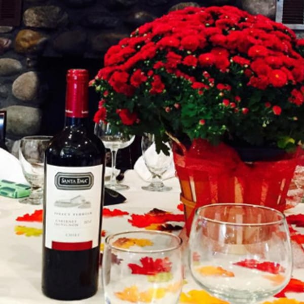 A bottle of Santa Emiliana wine, wine glasses, and a red mum centerpiece on a white tablecloth.